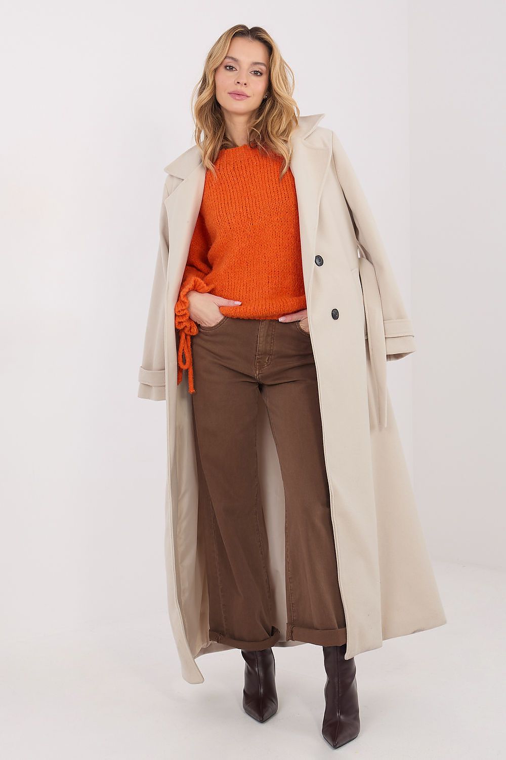 Woman wearing an orange sweater, brown pants, and a beige coat on a white background