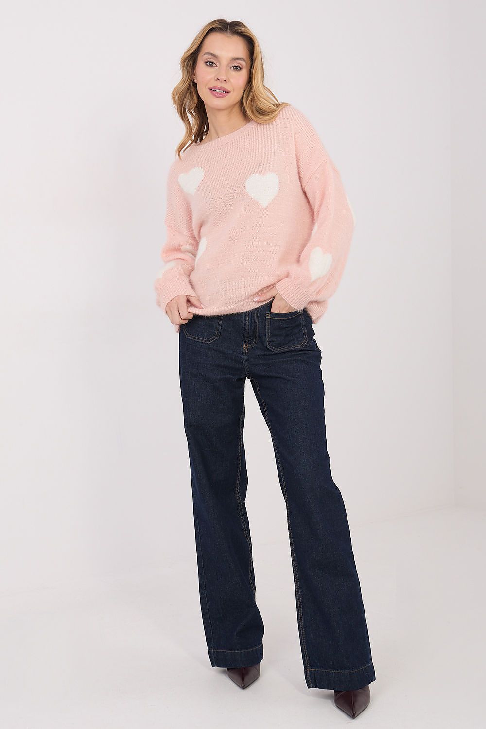 Woman wearing a pink sweater with white heart patterns and dark blue jeans on a white background