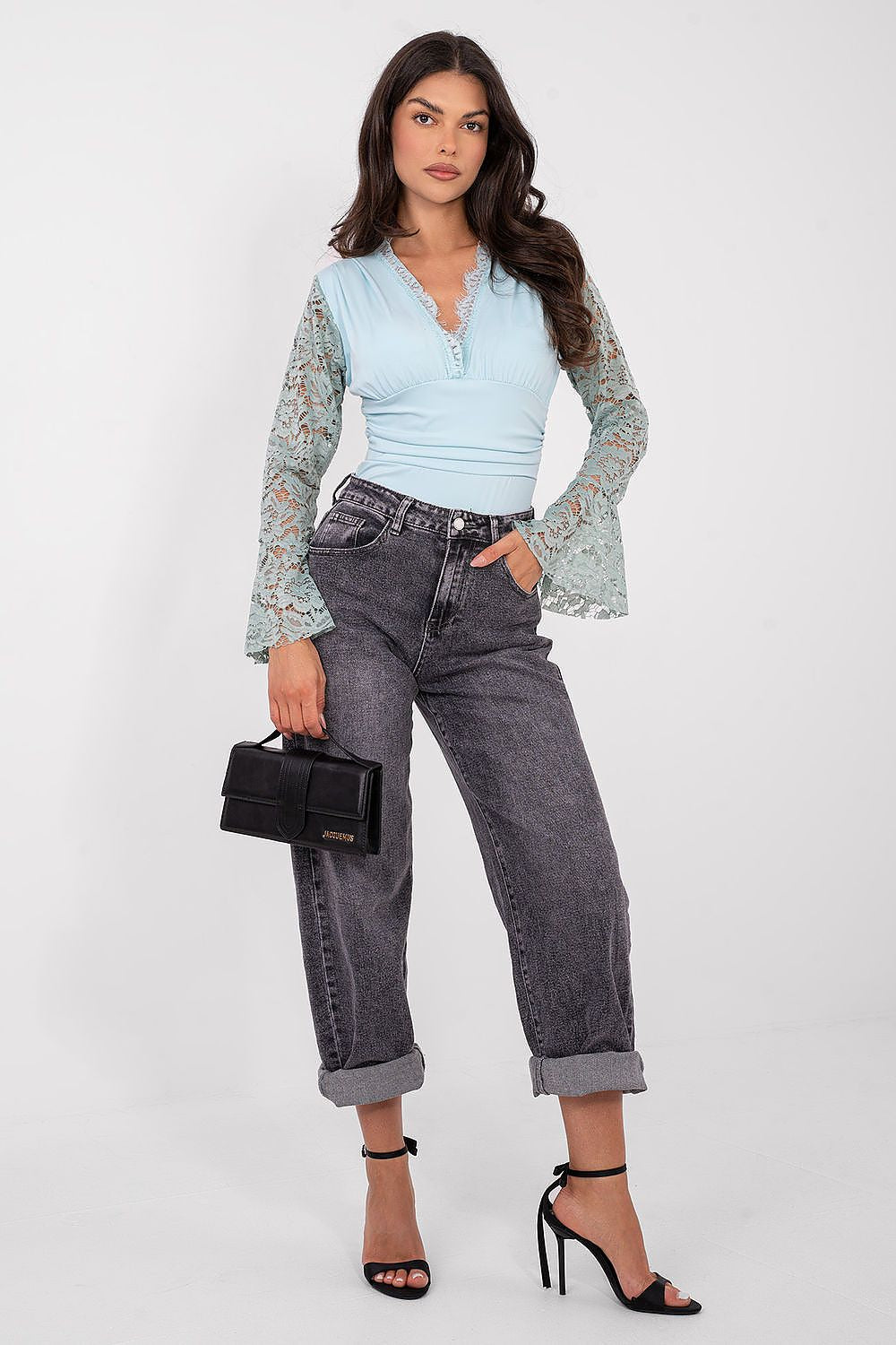 Woman wearing a light blue top and dark gray jeans, holding a black handbag on a white background