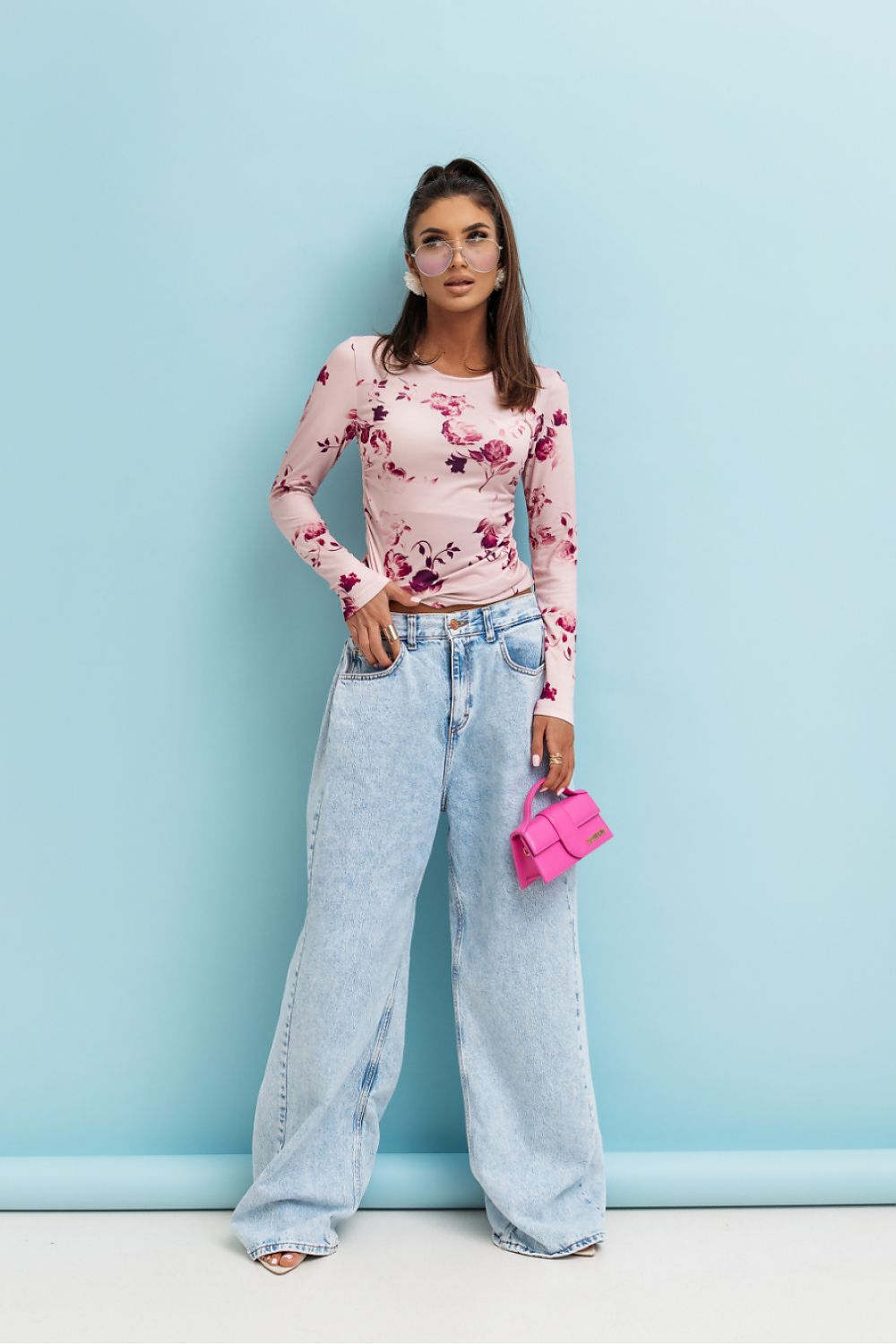 Woman wearing a floral top and jeans holding a pink clutch against a light blue background