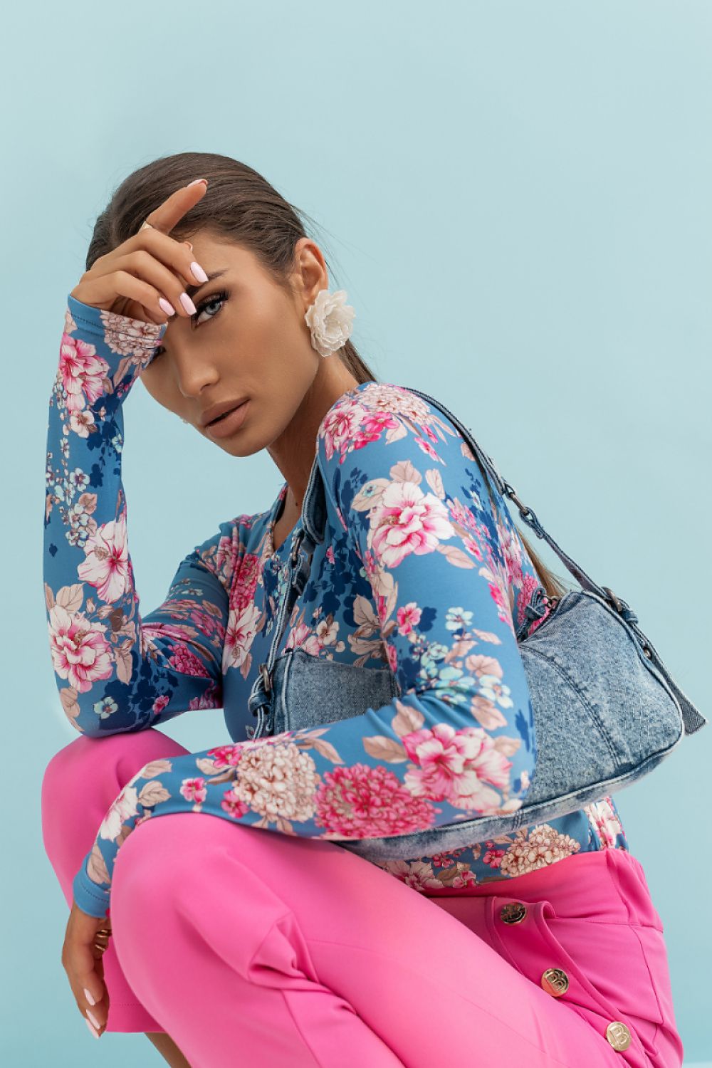 Woman wearing a floral jacket and pink pants, holding a gray handbag against a light blue background