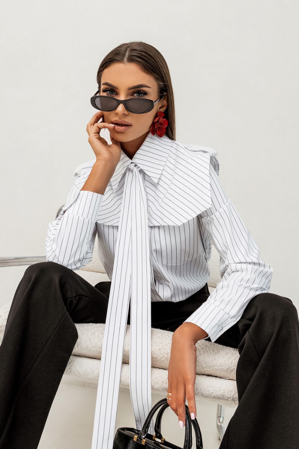 Woman wearing a white striped shirt with a large bow tie, black pants, and sunglasses on a white background.