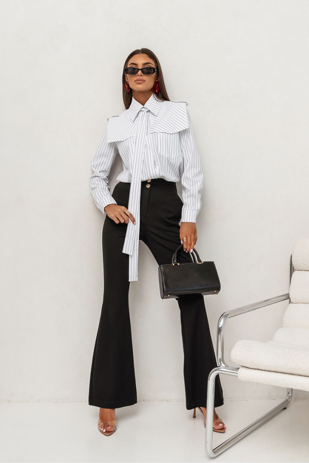 Woman in a white shirt with a large collar and black pants holding a black handbag against a white background.