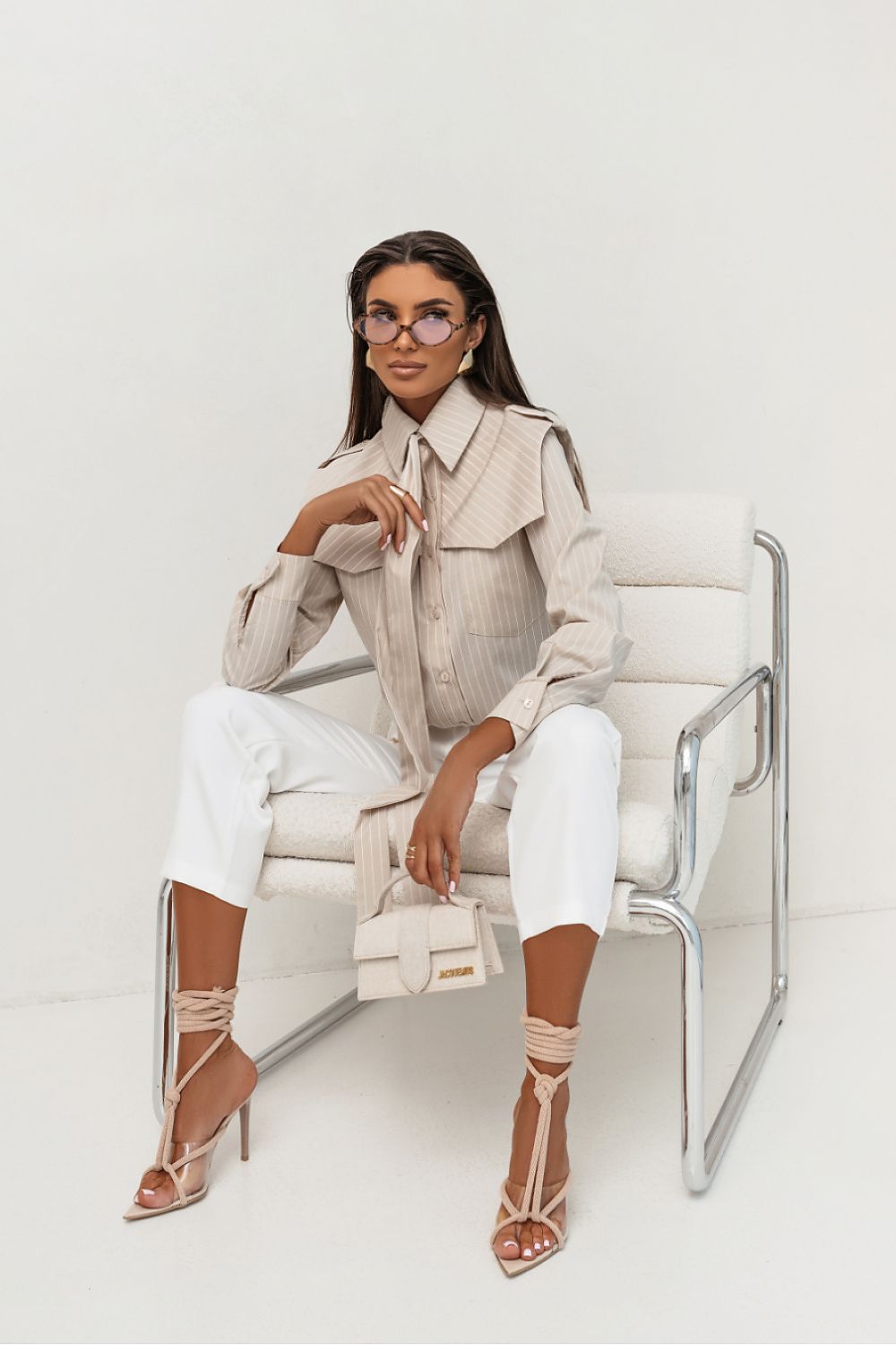 Woman sitting on a white chair wearing a beige outfit with a white background
