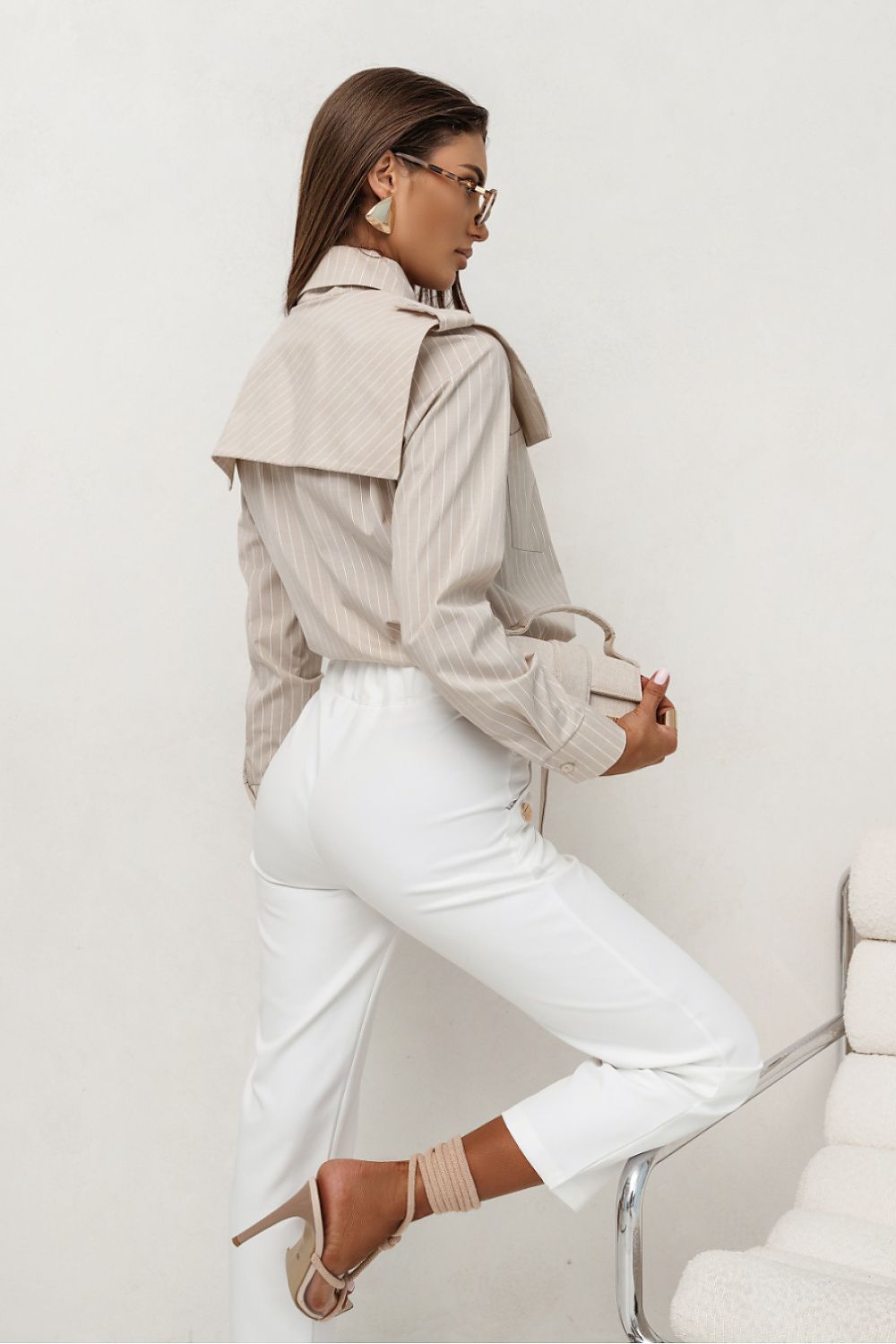 Woman wearing a beige trench coat over a white shirt and pants, standing against a white background.