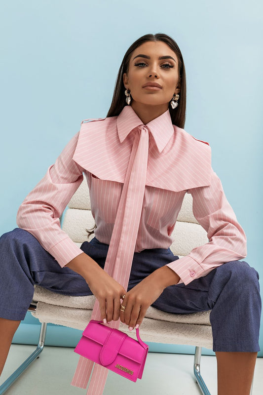 Woman wearing a pink blouse with a large collar and holding a pink handbag against a light blue background.