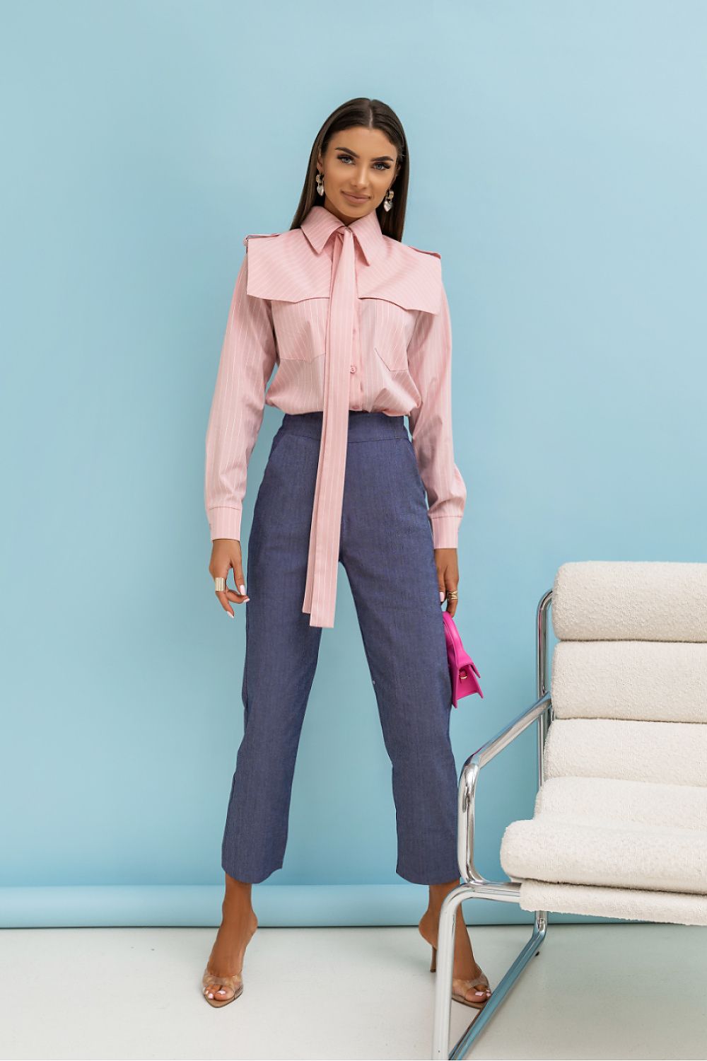 Woman wearing a pink blouse and blue pants against a light blue wall with a chair.