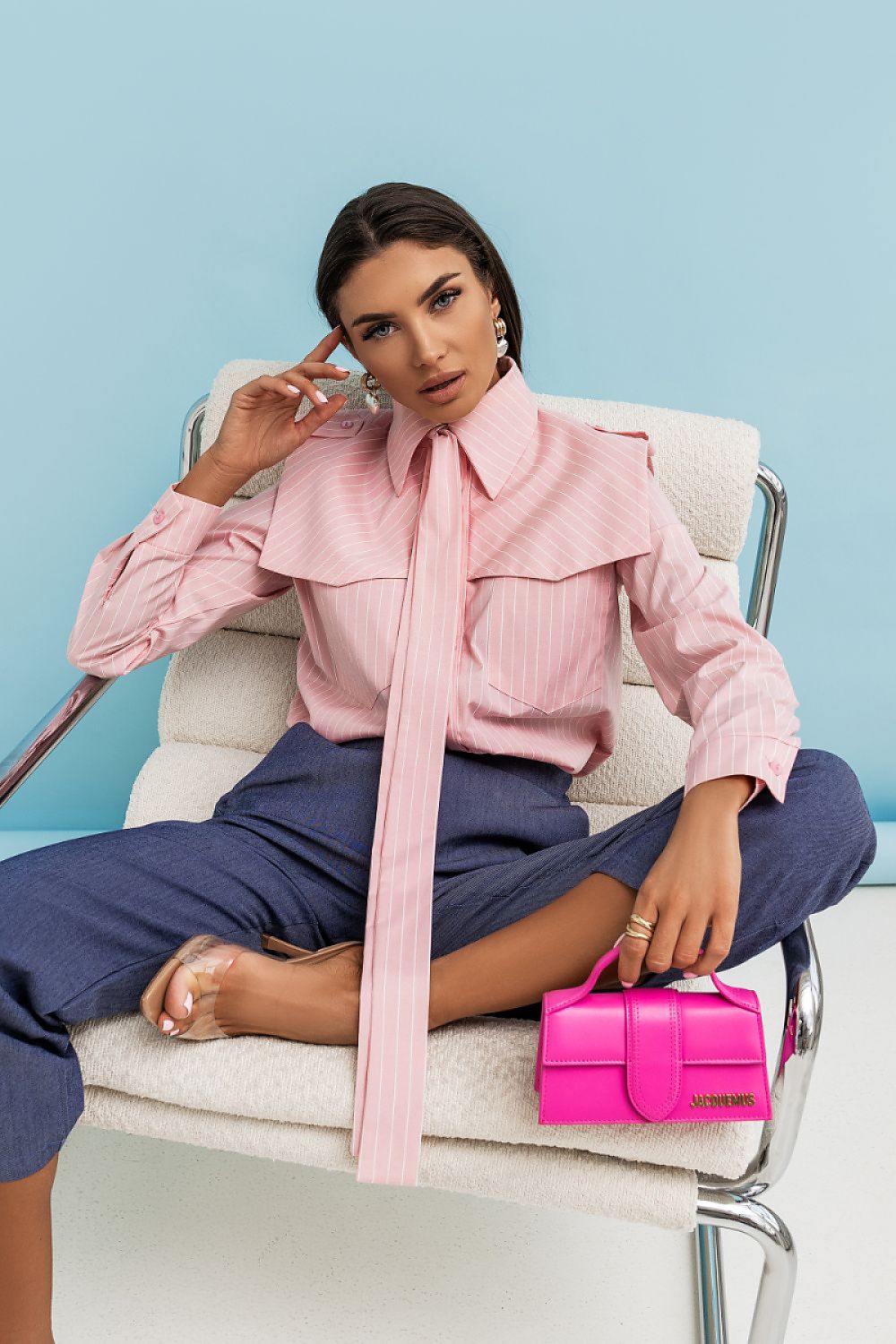 Woman in a pink blouse with a large bow sitting on a chair holding a pink handbag against a light blue background.