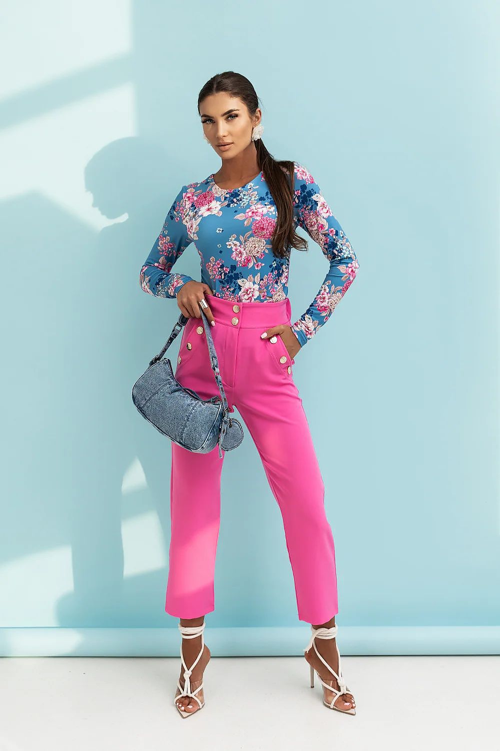 Woman in floral top and pink pants holding a blue handbag against a light blue wall.