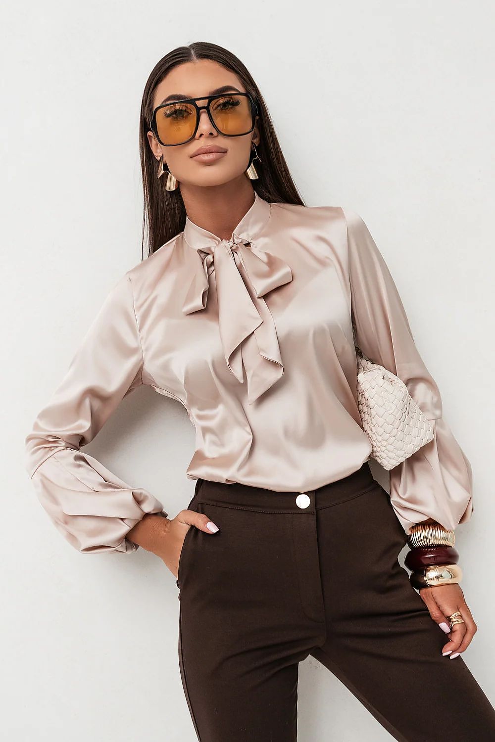 Woman wearing a beige satin blouse with ruffles and brown pants against a white background