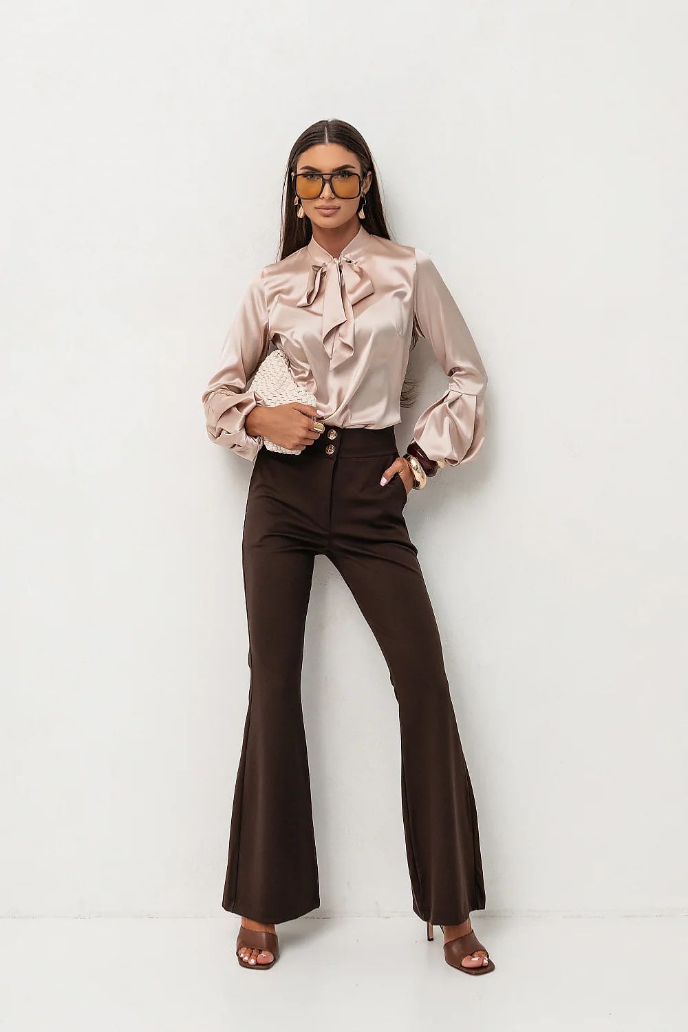 Woman wearing a beige blouse with a bow and brown flared pants on a white background