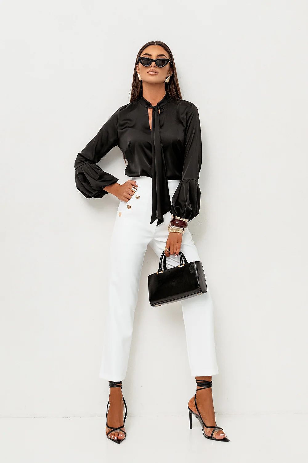 Woman wearing a black blouse, white pants, and black heels against a white background