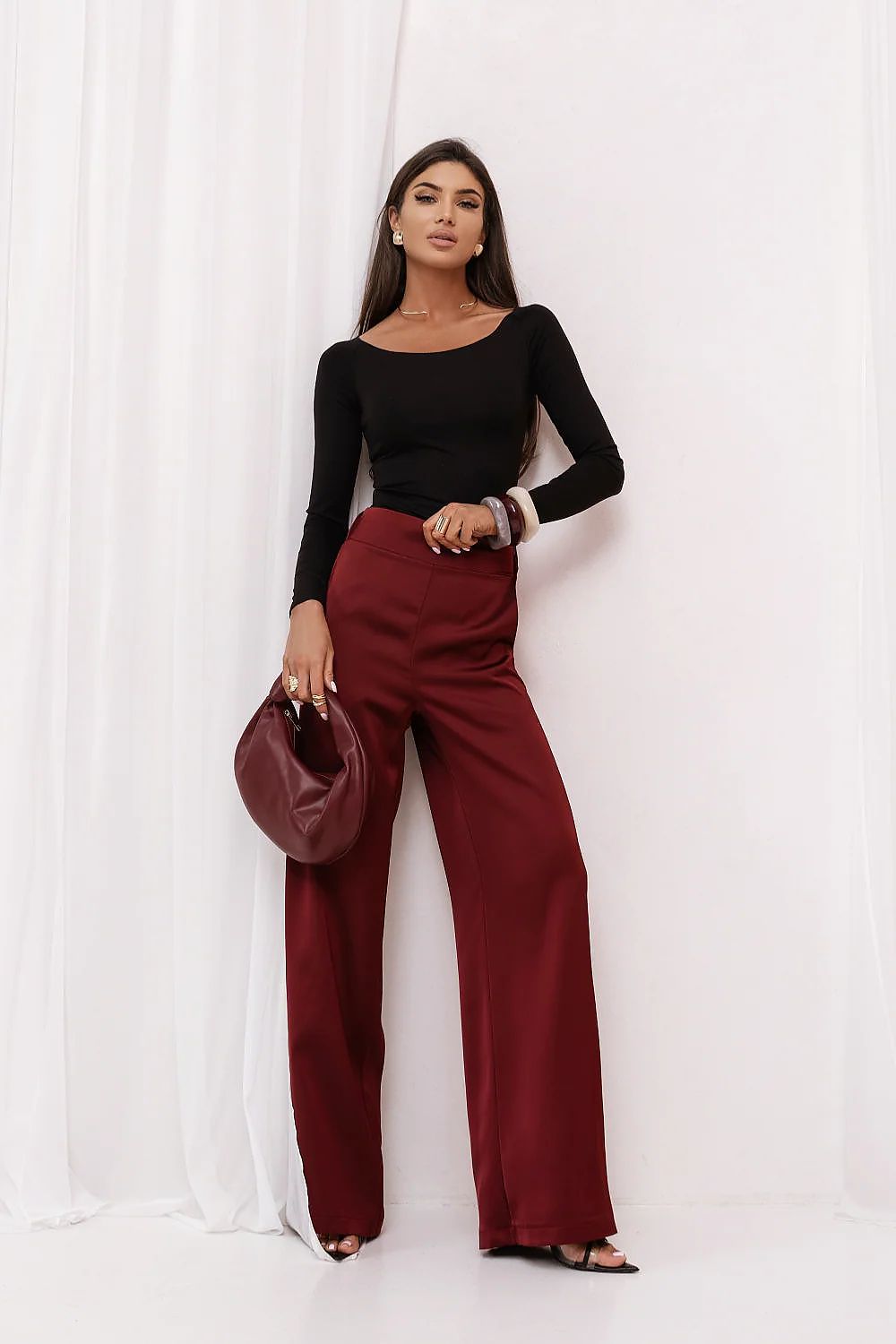 Woman wearing a black top and red pants holding a brown handbag against a white curtain background