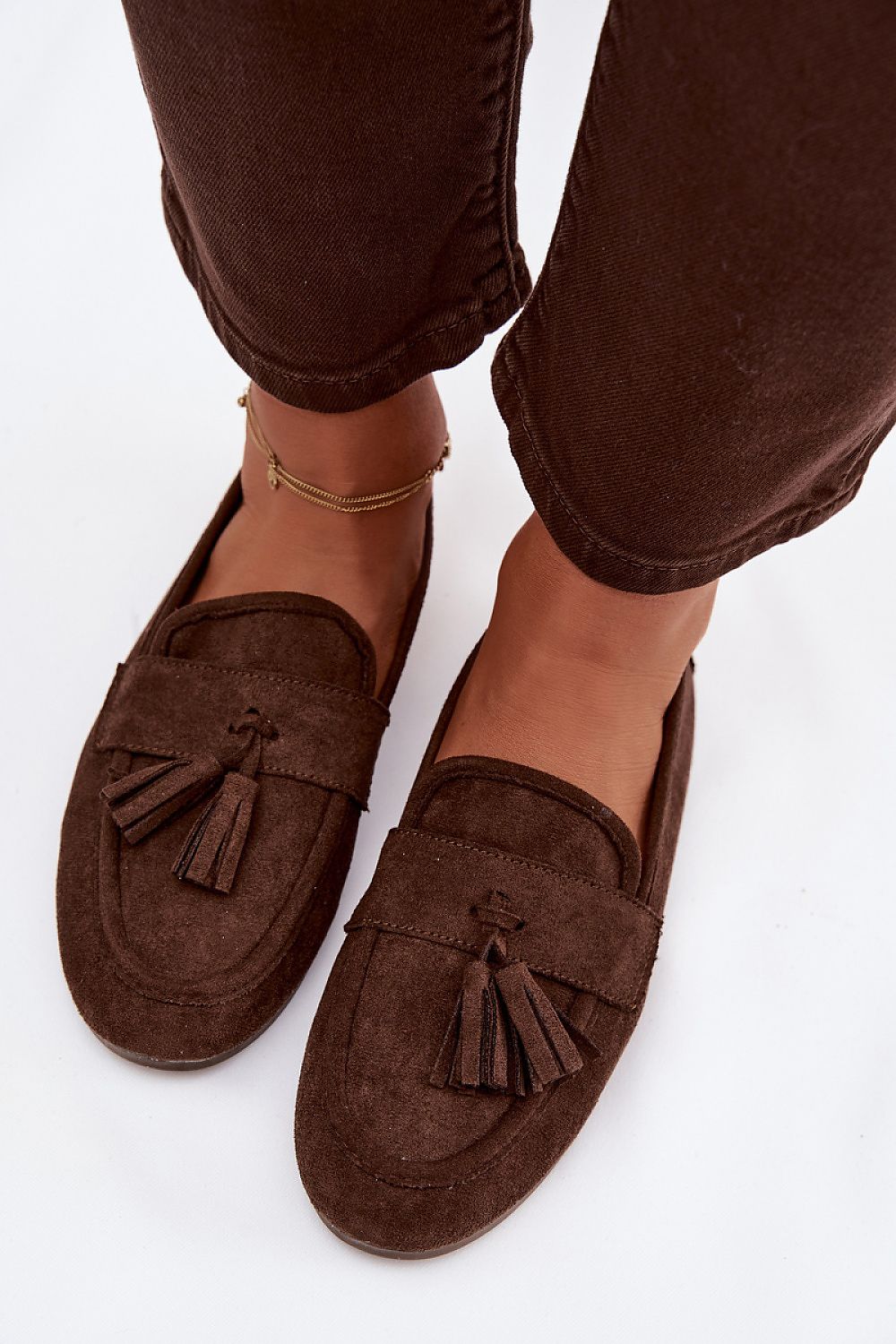 Brown suede loafers with tassels worn with brown pants on a white background