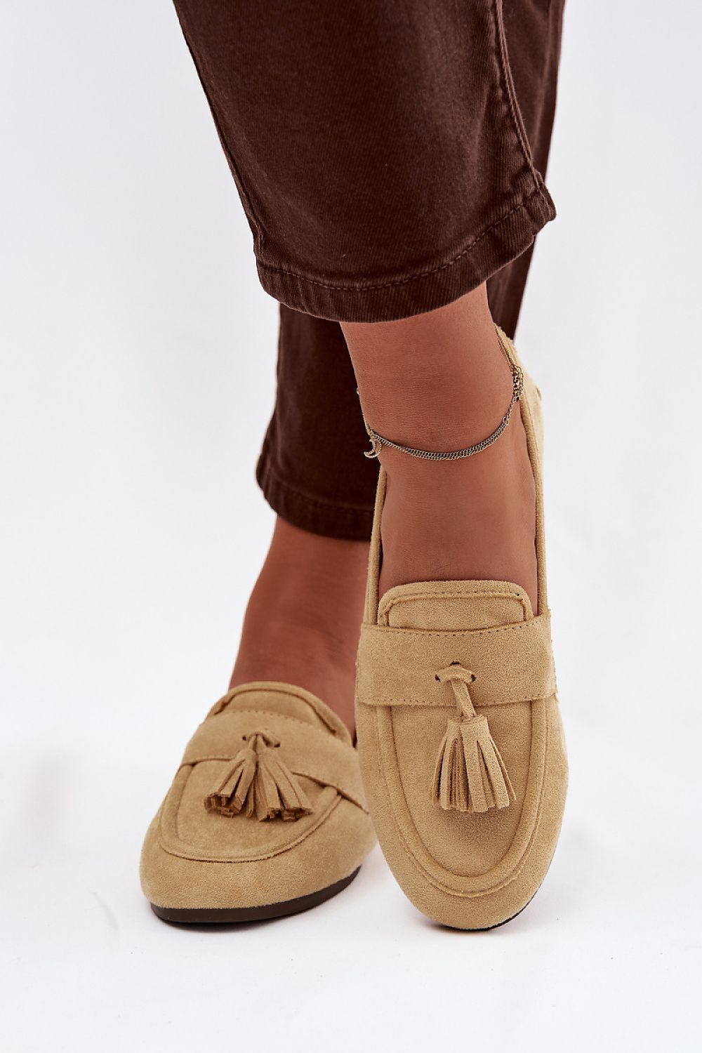 Beige loafers with tassels worn with brown pants on a white background