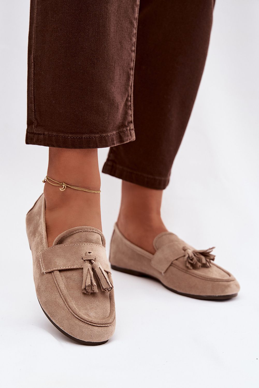 Beige loafers with tassels worn with brown pants on a white background