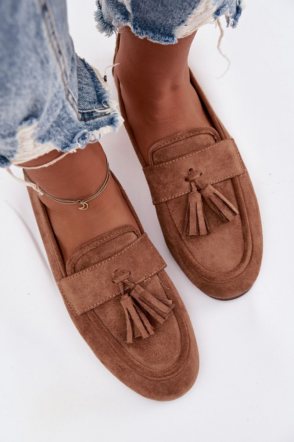 Brown loafers with tassels worn with ripped jeans on a white background