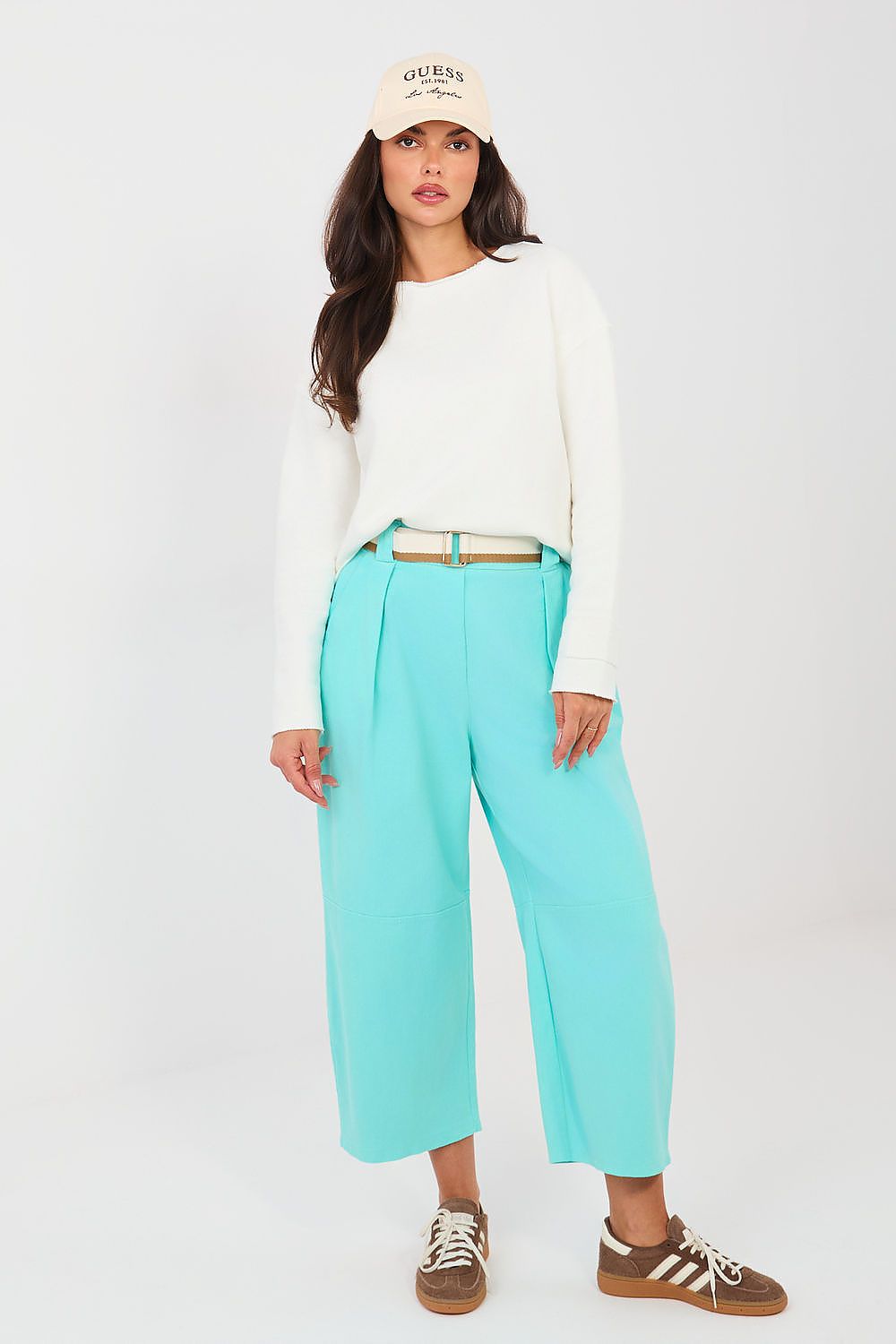 Woman wearing a white long-sleeve top, turquoise pants, and a beige cap on a white background