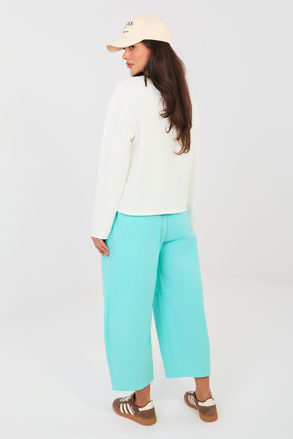Woman wearing a white top and turquoise pants with a beige cap on a white background