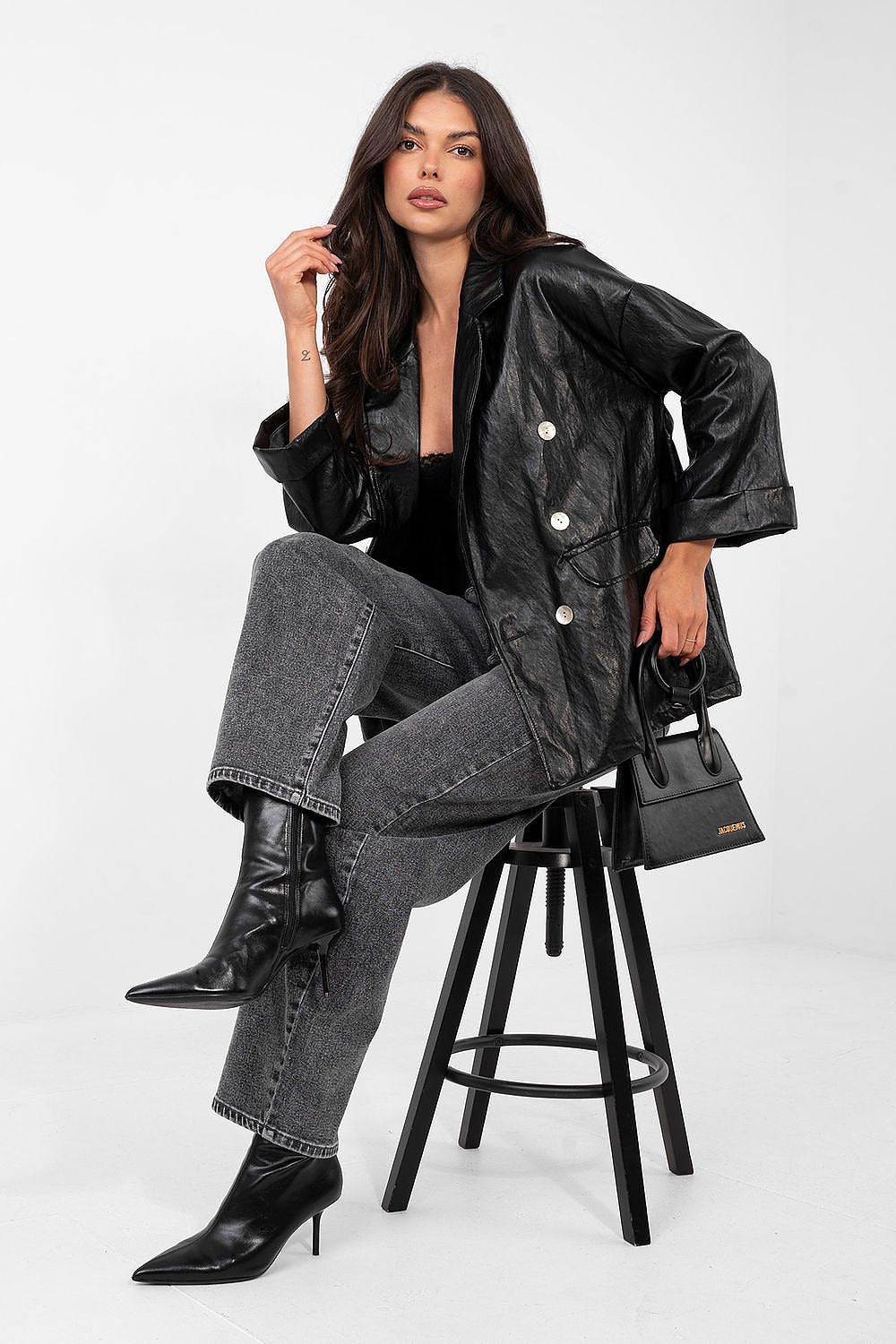 Woman sitting on a stool wearing a black leather jacket, black jeans, and black boots, holding a black handbag.