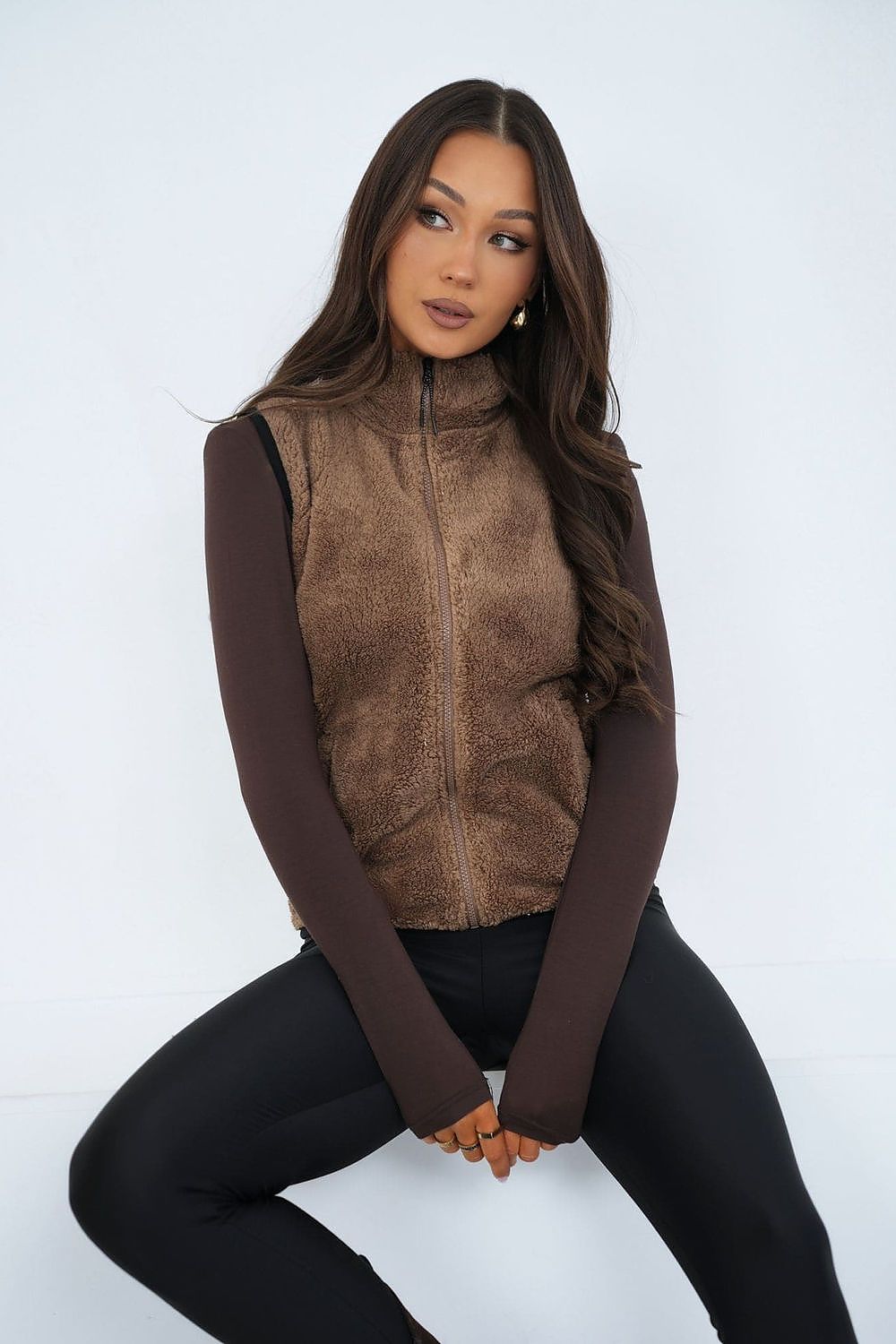 Woman wearing a brown fleece vest over a long-sleeve shirt and black pants on a white background