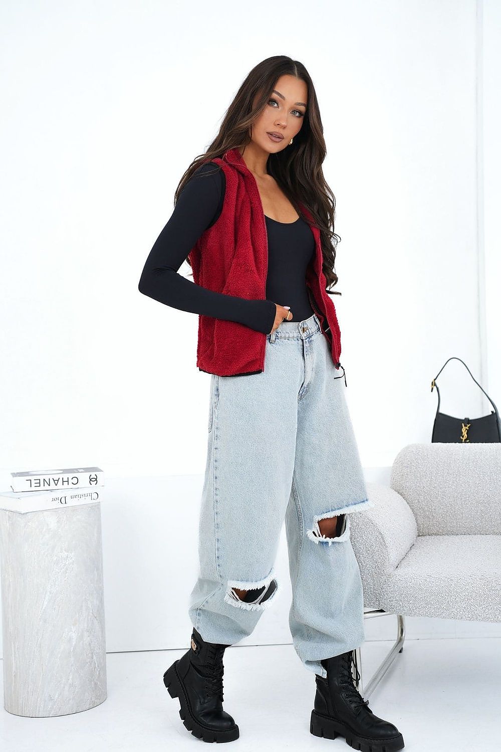 Woman wearing a red vest, black top, light blue jeans, and black boots in a minimalistic room.