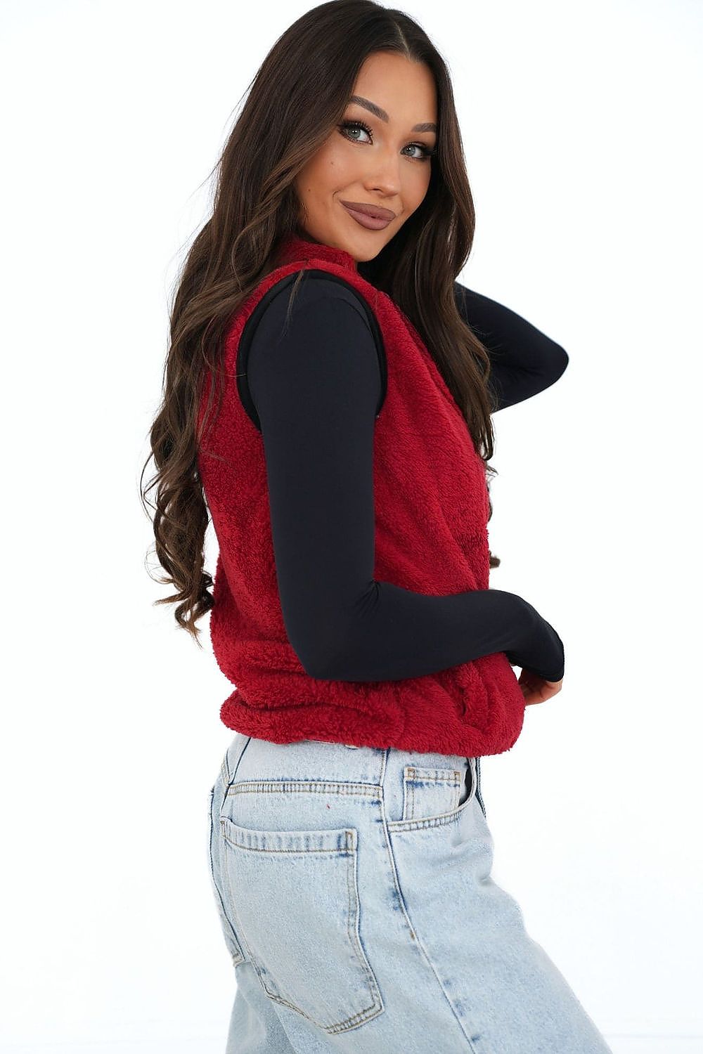 Woman wearing a red vest over a black long-sleeve shirt and light blue jeans on a white background