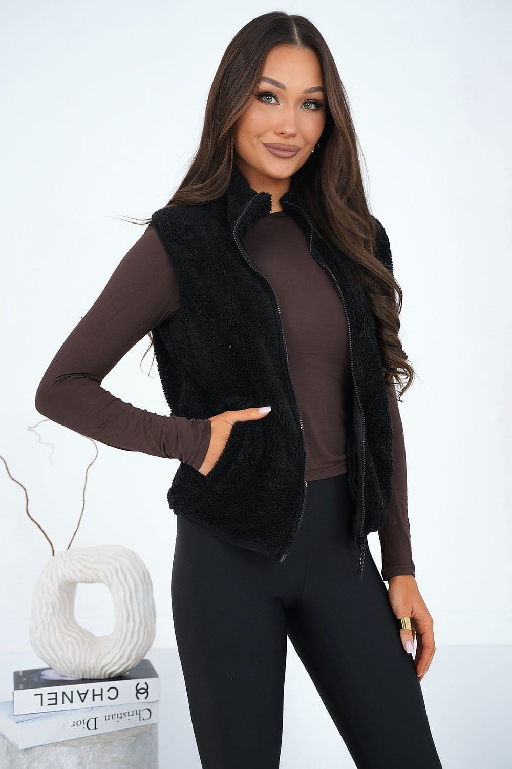 Woman wearing a black fur vest over a brown top and black leggings, standing against a white background.