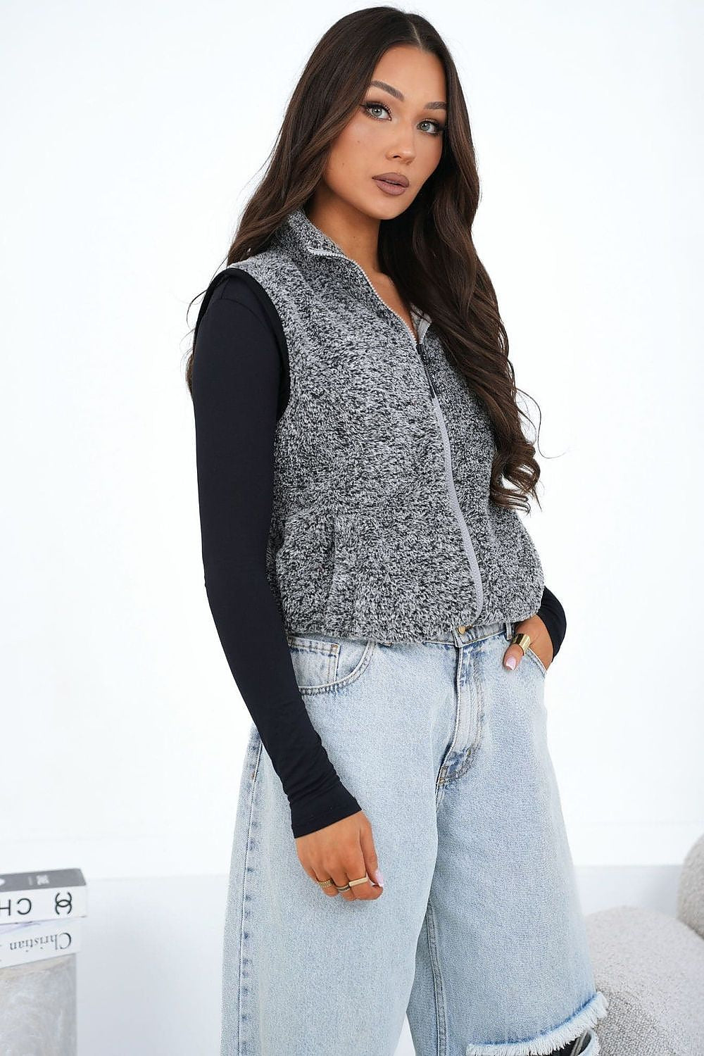 Woman wearing a gray marled vest over a black long-sleeve shirt and light blue jeans on a white background.