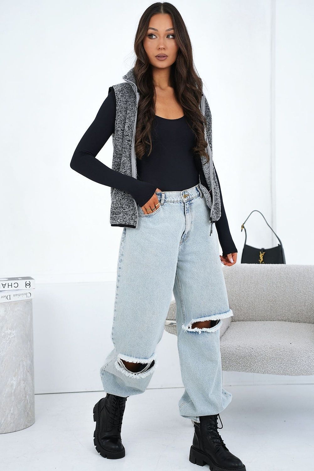 Woman wearing a black top, gray vest, light blue jeans, and black boots in a minimalistic room.
