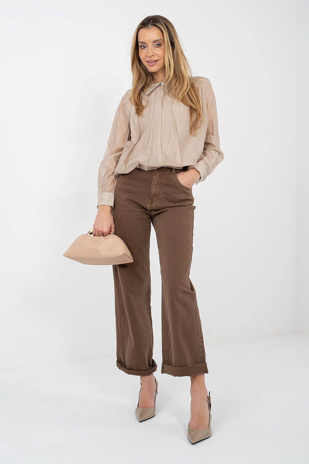 Woman wearing a beige blouse and brown pants holding a beige handbag on a white background