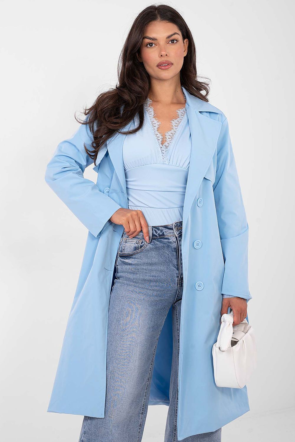 Woman wearing a light blue coat over a light blue top and jeans, holding a white handbag on a white background.