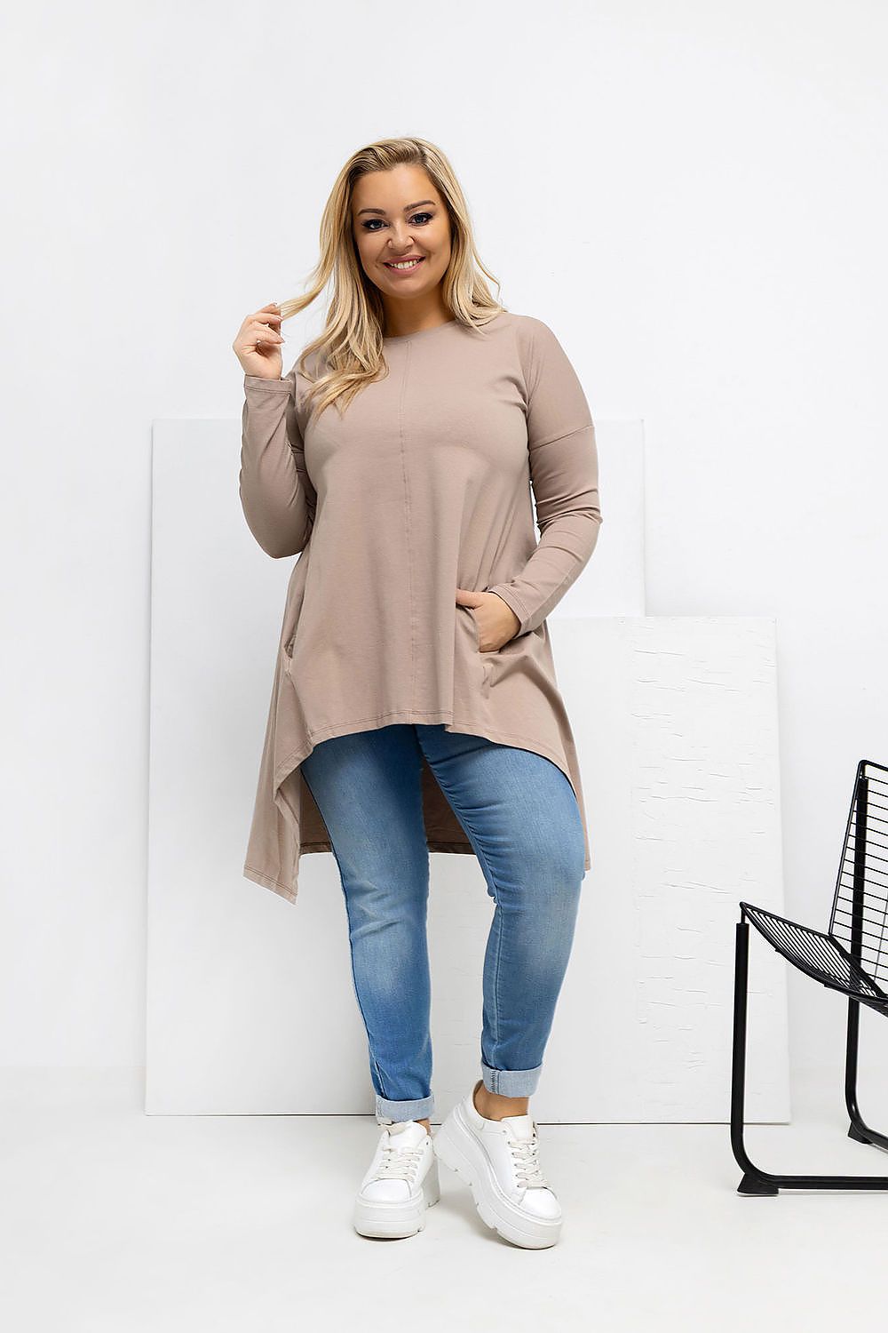 Woman wearing a beige long-sleeve top and blue jeans standing against a white background.