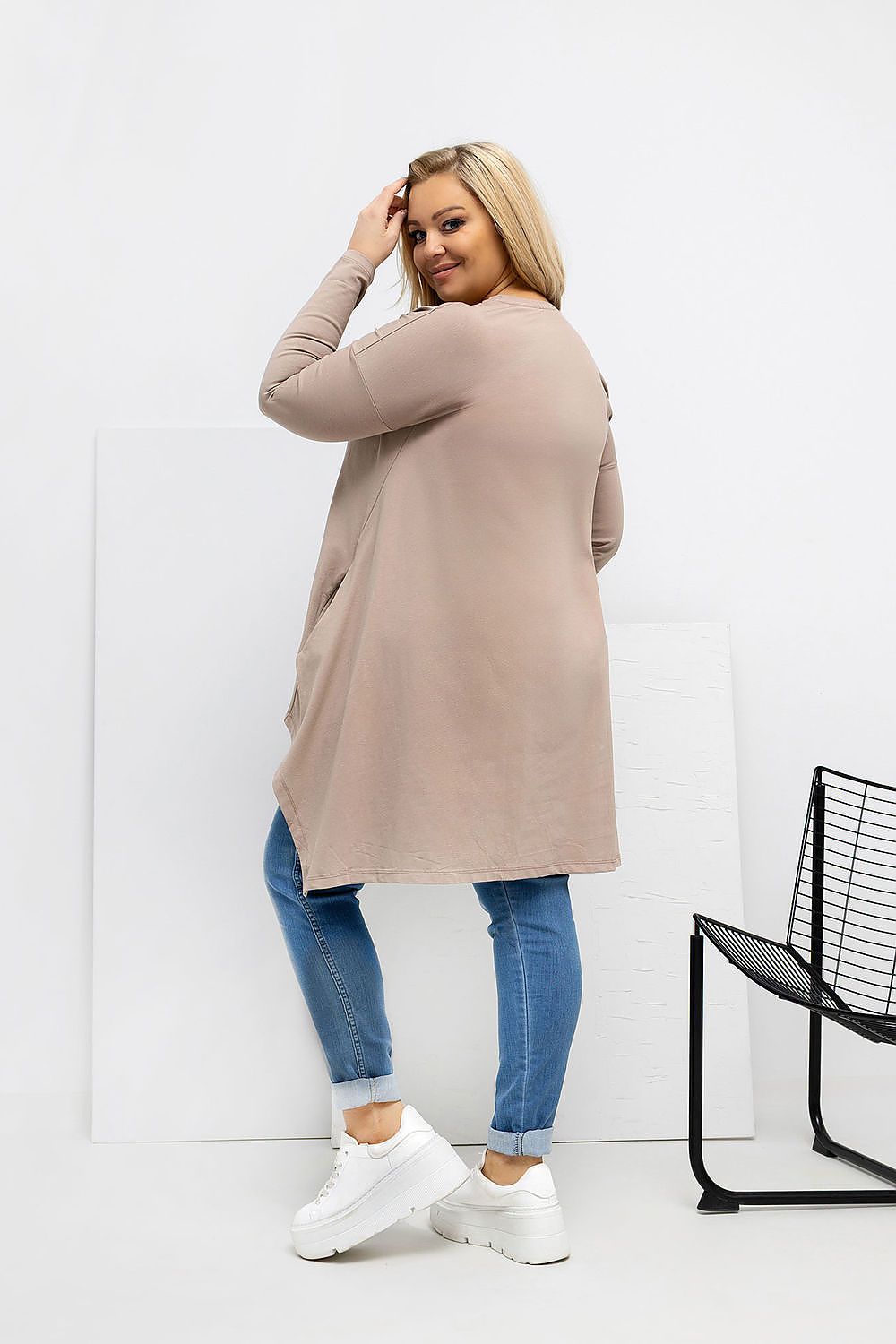 Woman wearing a beige cardigan, blue jeans, and white sneakers on a white background