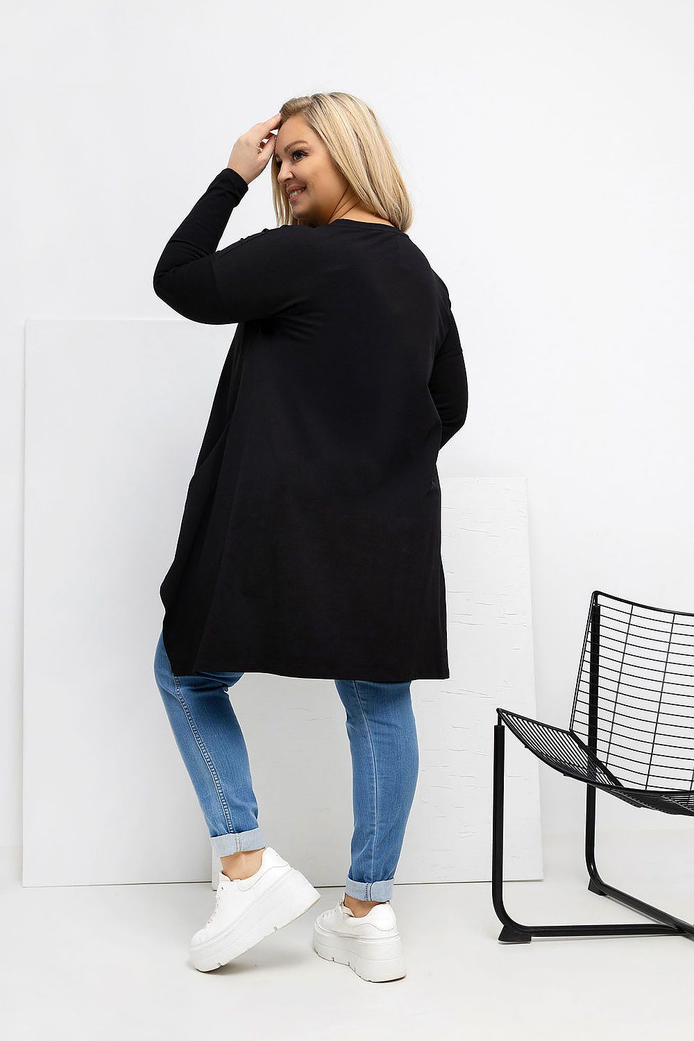 Woman wearing a black cardigan, blue jeans, and white sneakers against a white background.