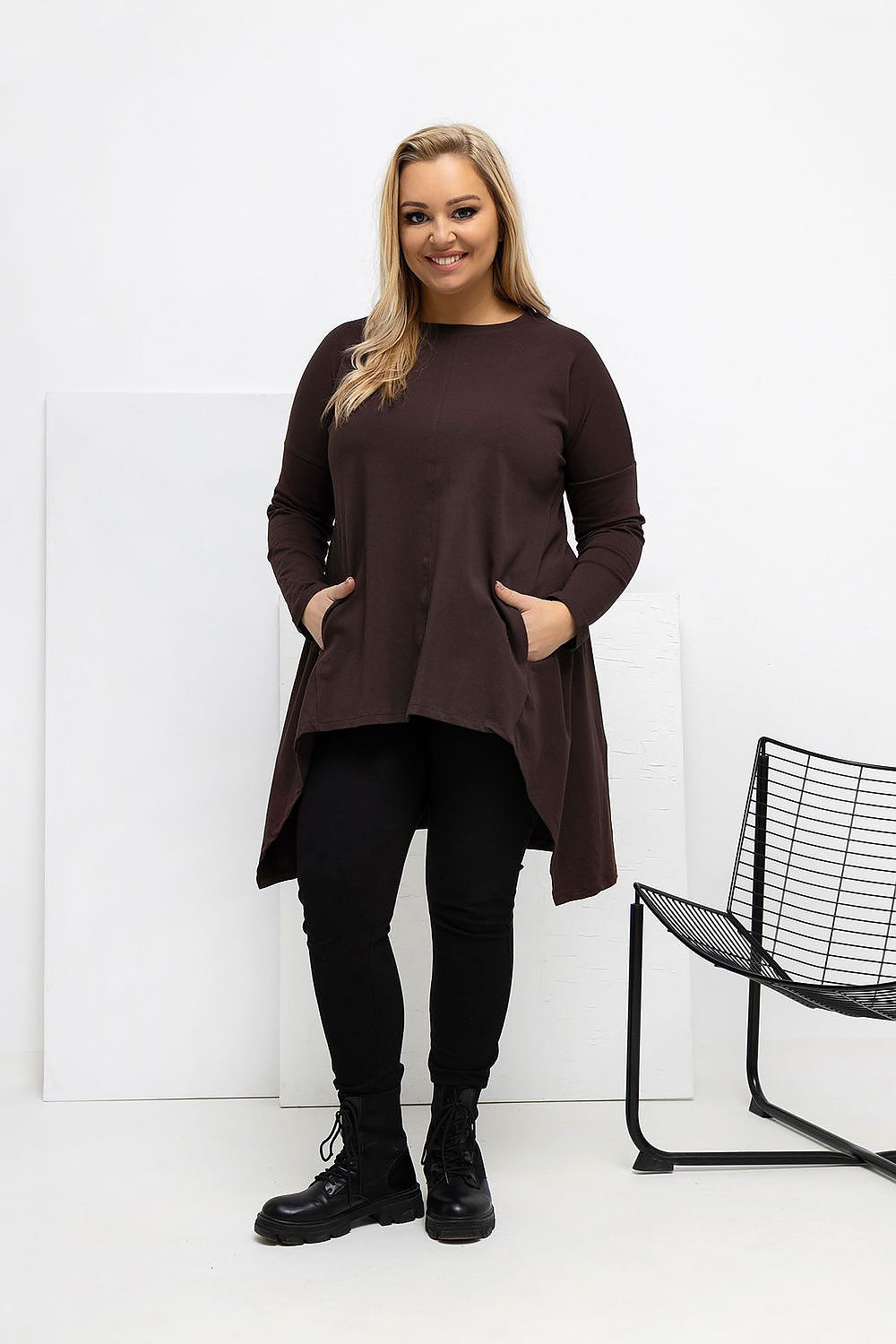 Woman wearing a brown dress and black pants standing in a minimalistic room with a black chair.
