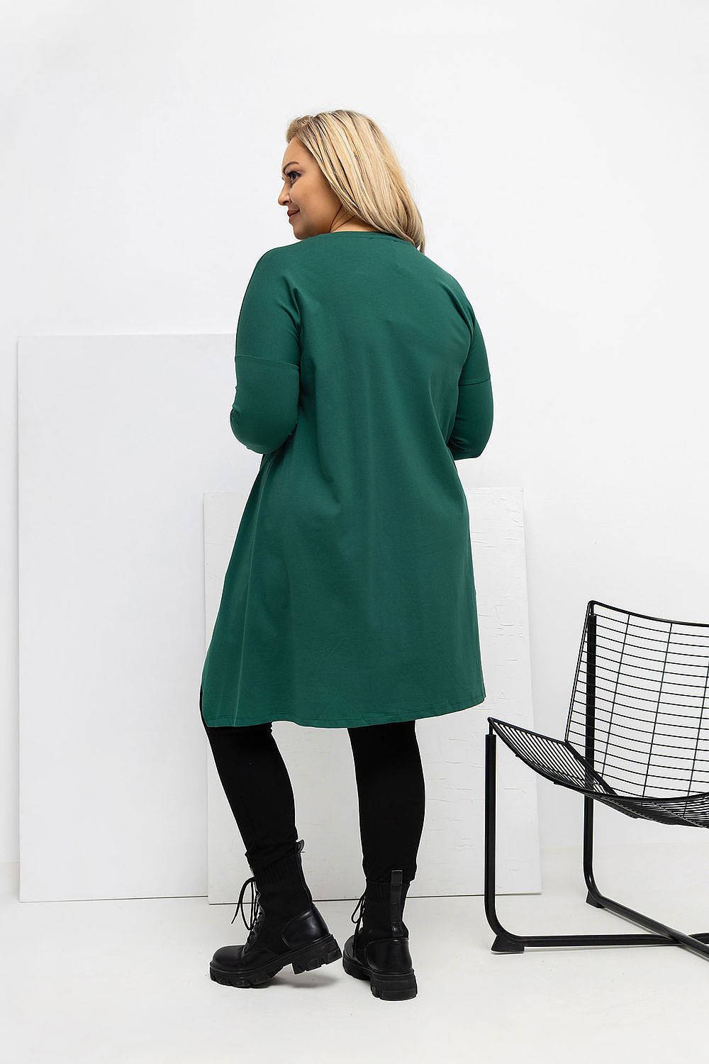 Woman wearing a green coat standing against a white wall with a black chair.