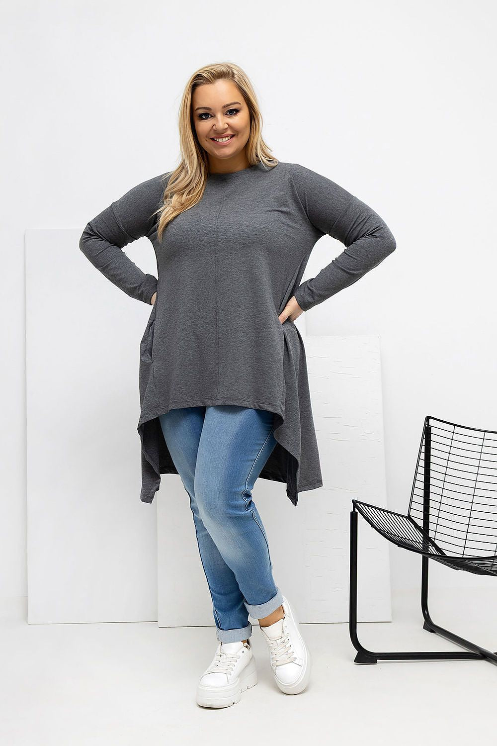 Woman wearing a long gray sweater and blue jeans standing against a white background.