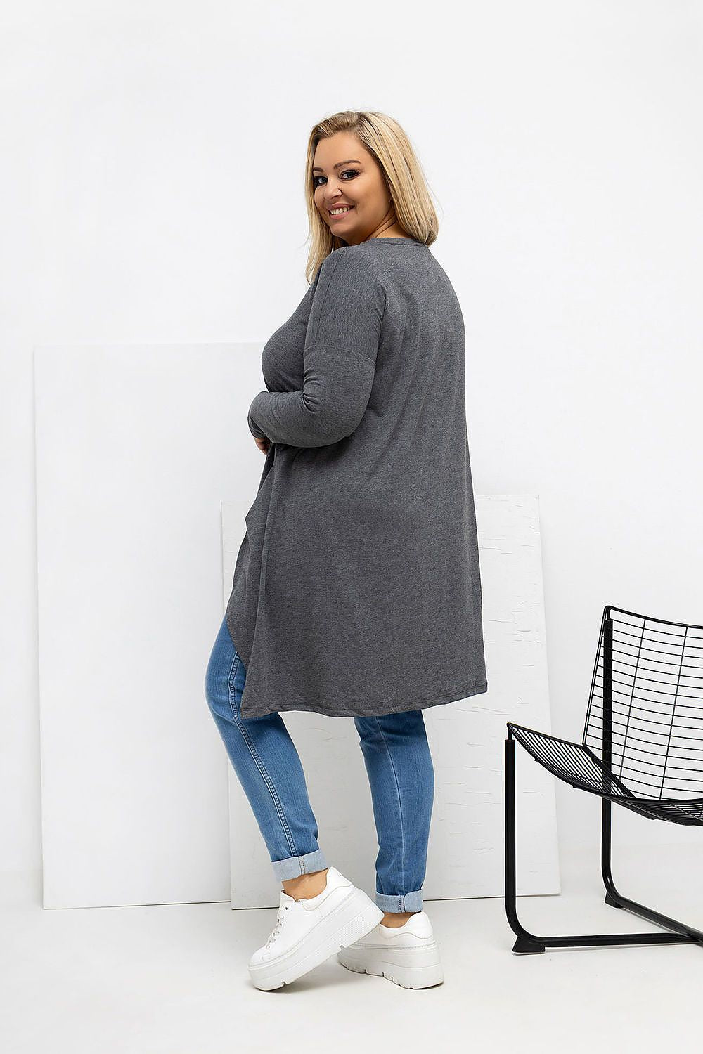 Woman wearing a long gray cardigan over blue jeans with white sneakers, standing against a white background.