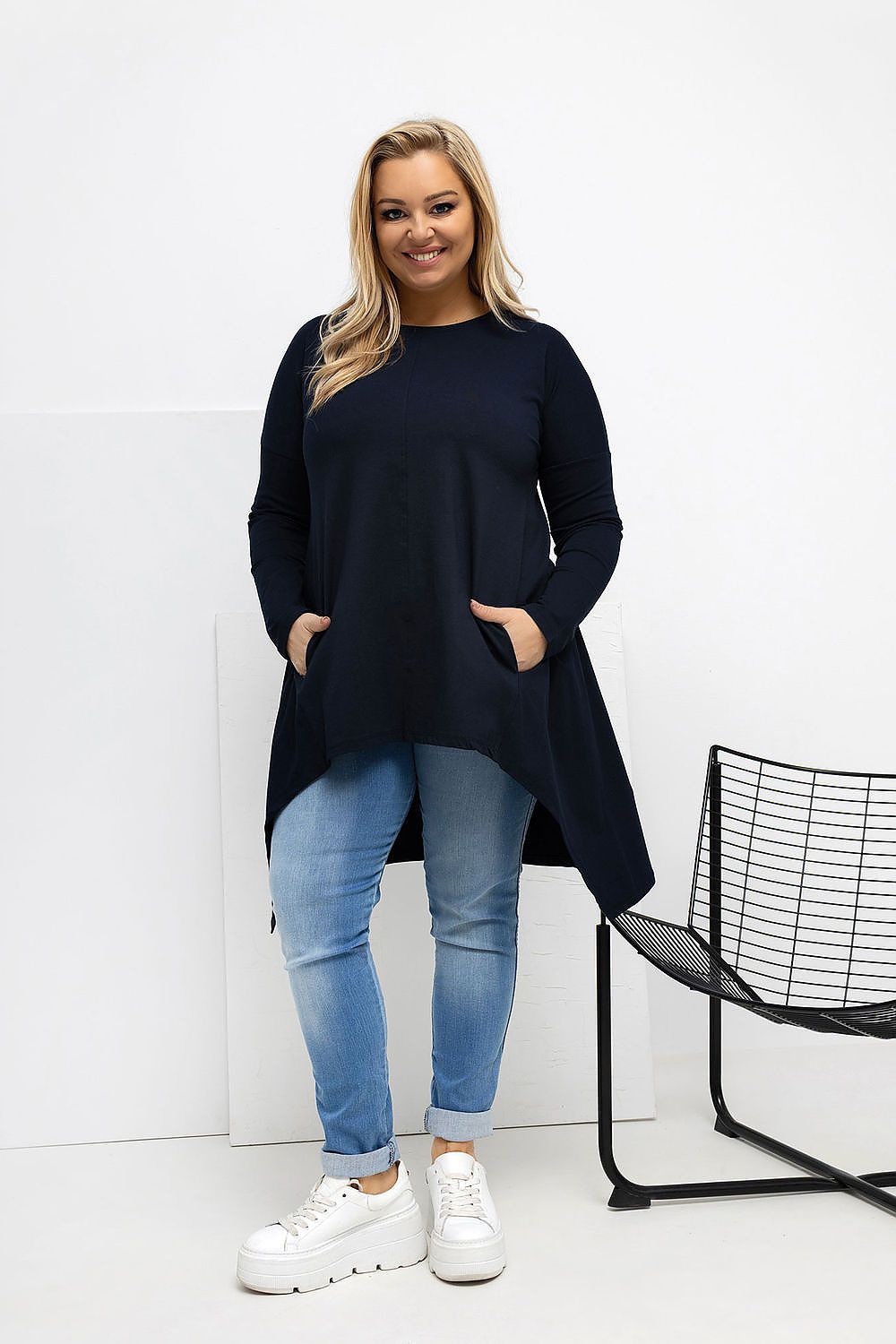 Woman wearing a black long-sleeve top and blue jeans standing against a white background.