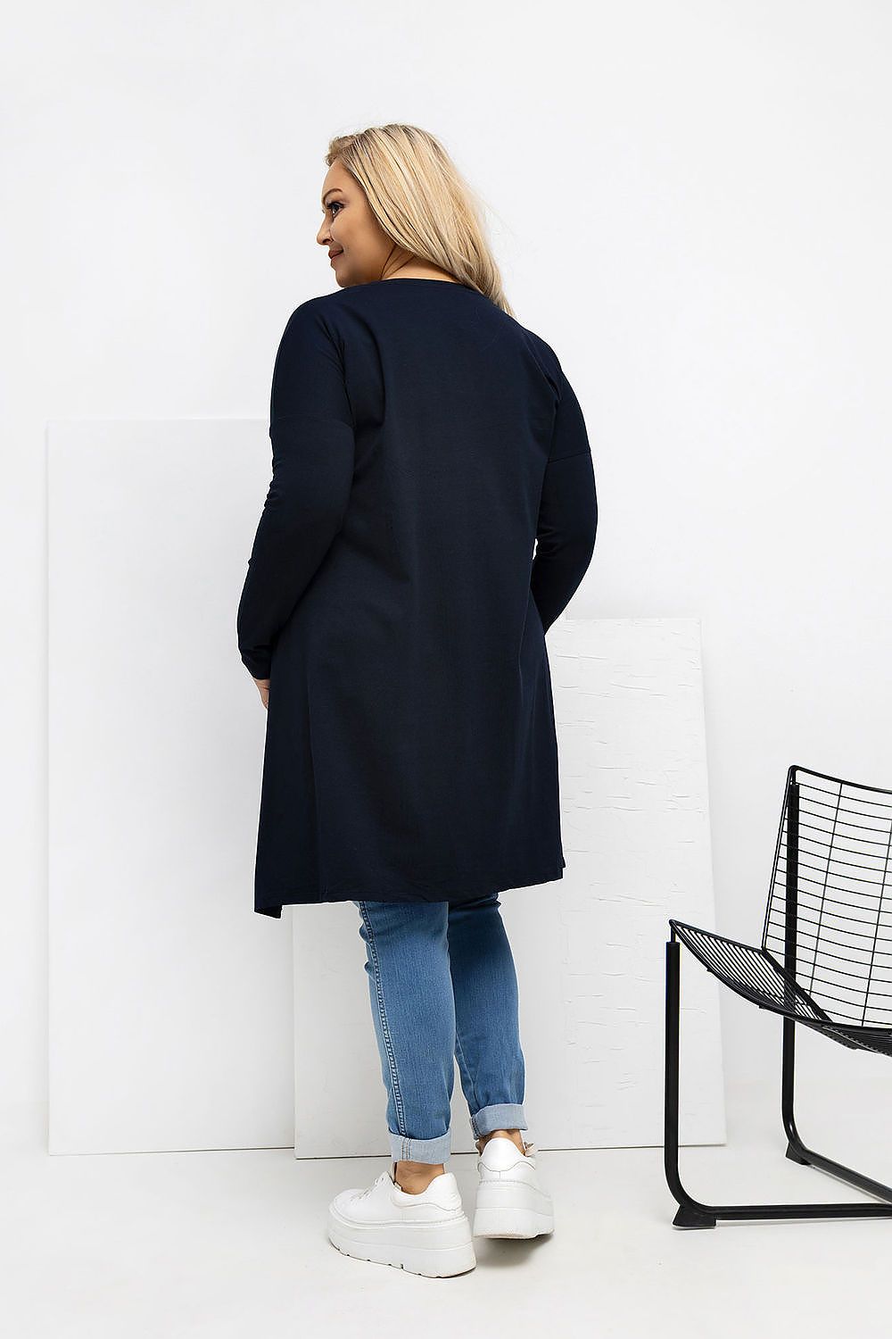 Woman wearing a navy coat standing against a white wall with a black chair.