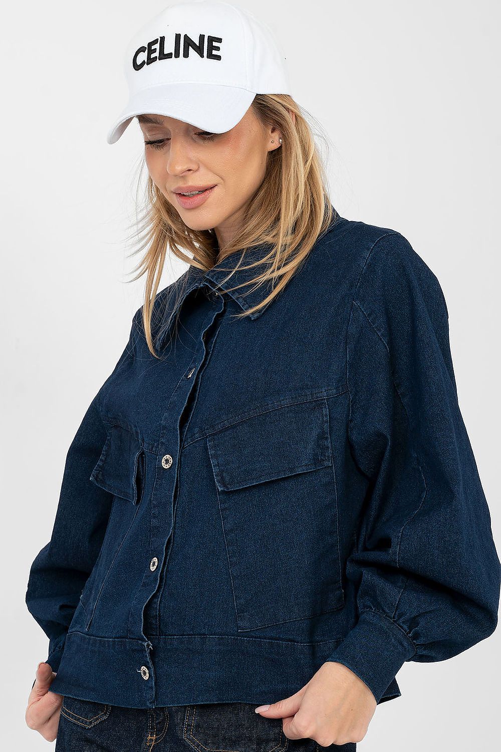 Person wearing a navy blue denim jacket and a white Celine cap on a plain background