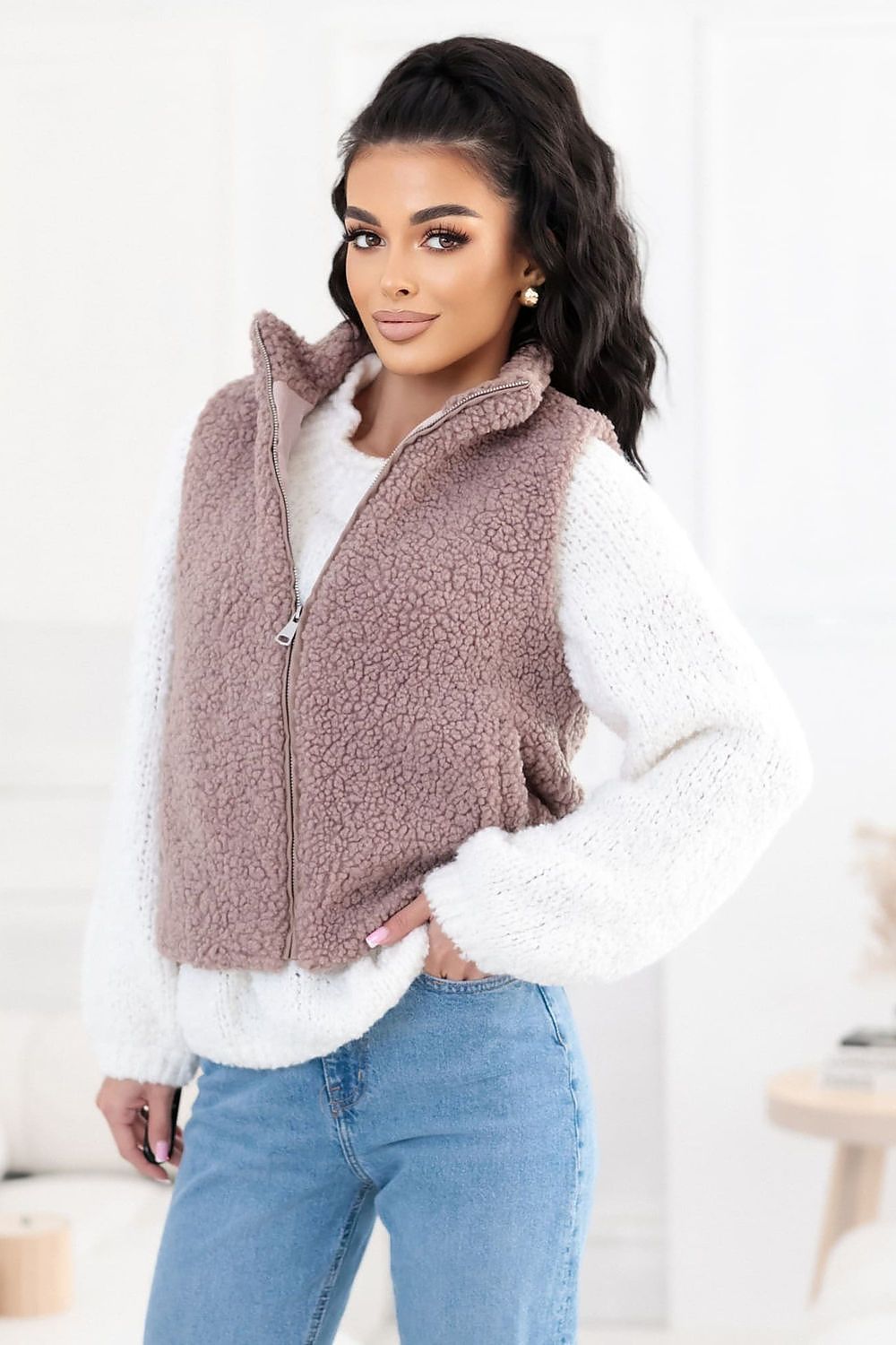 Woman wearing a brown fleece vest over a white shirt with blue jeans indoors.