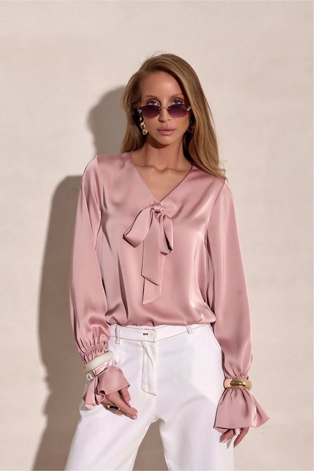 Woman wearing a pink blouse with a tie detail and white pants against a beige background