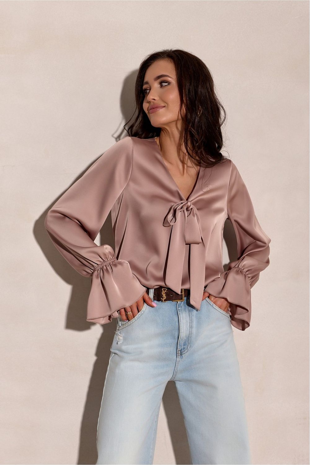 Woman wearing a pink blouse with long sleeves and light blue jeans against a beige background