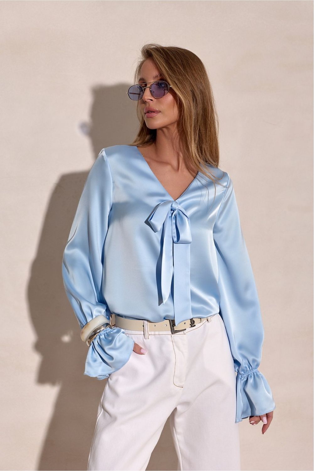 Woman wearing a light blue blouse with a tie detail and white pants against a beige background