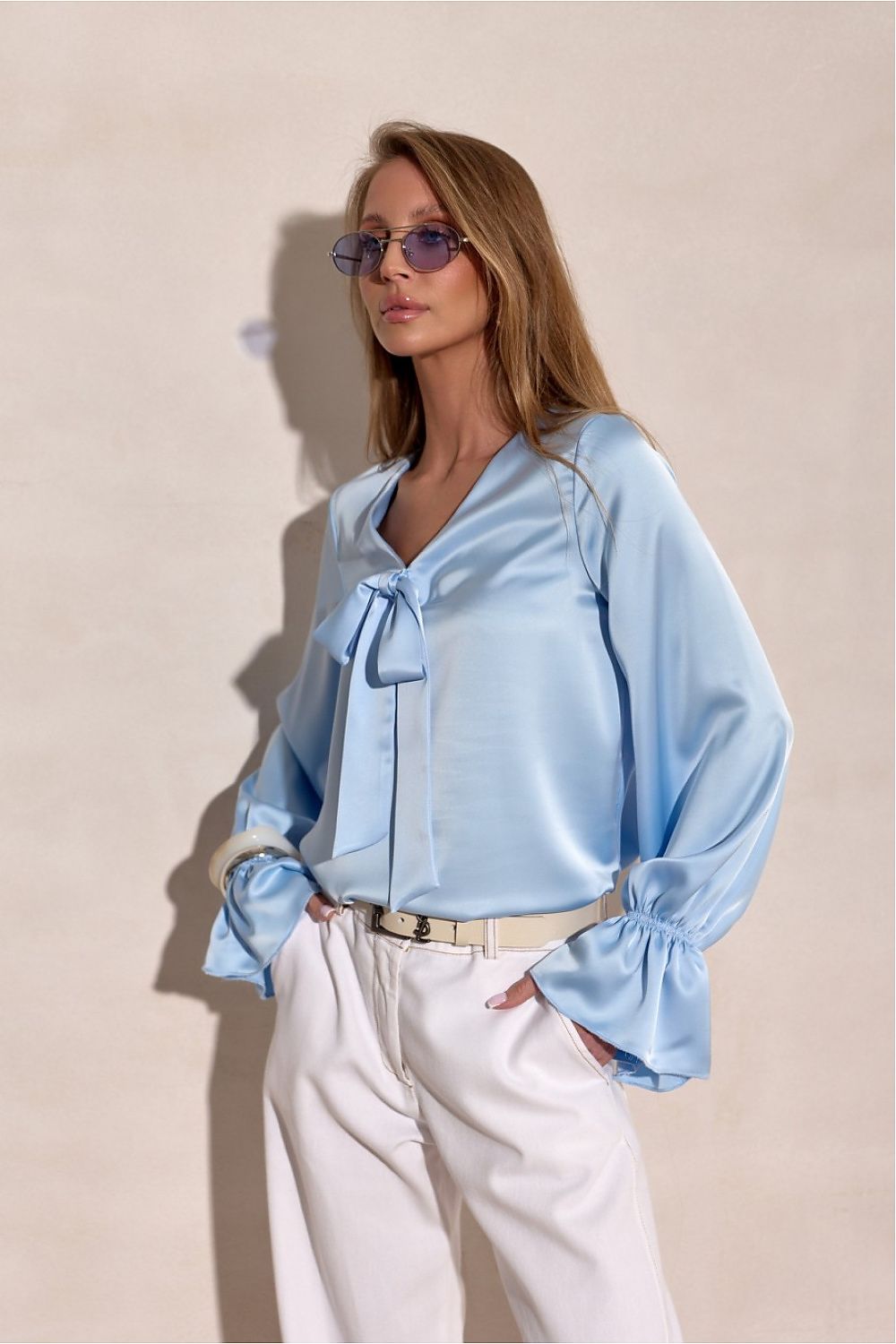 Woman wearing a light blue blouse with a bow tie and white pants against a beige background