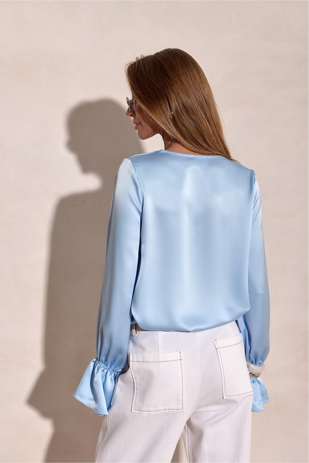 Woman wearing a light blue blouse and white pants against a beige wall