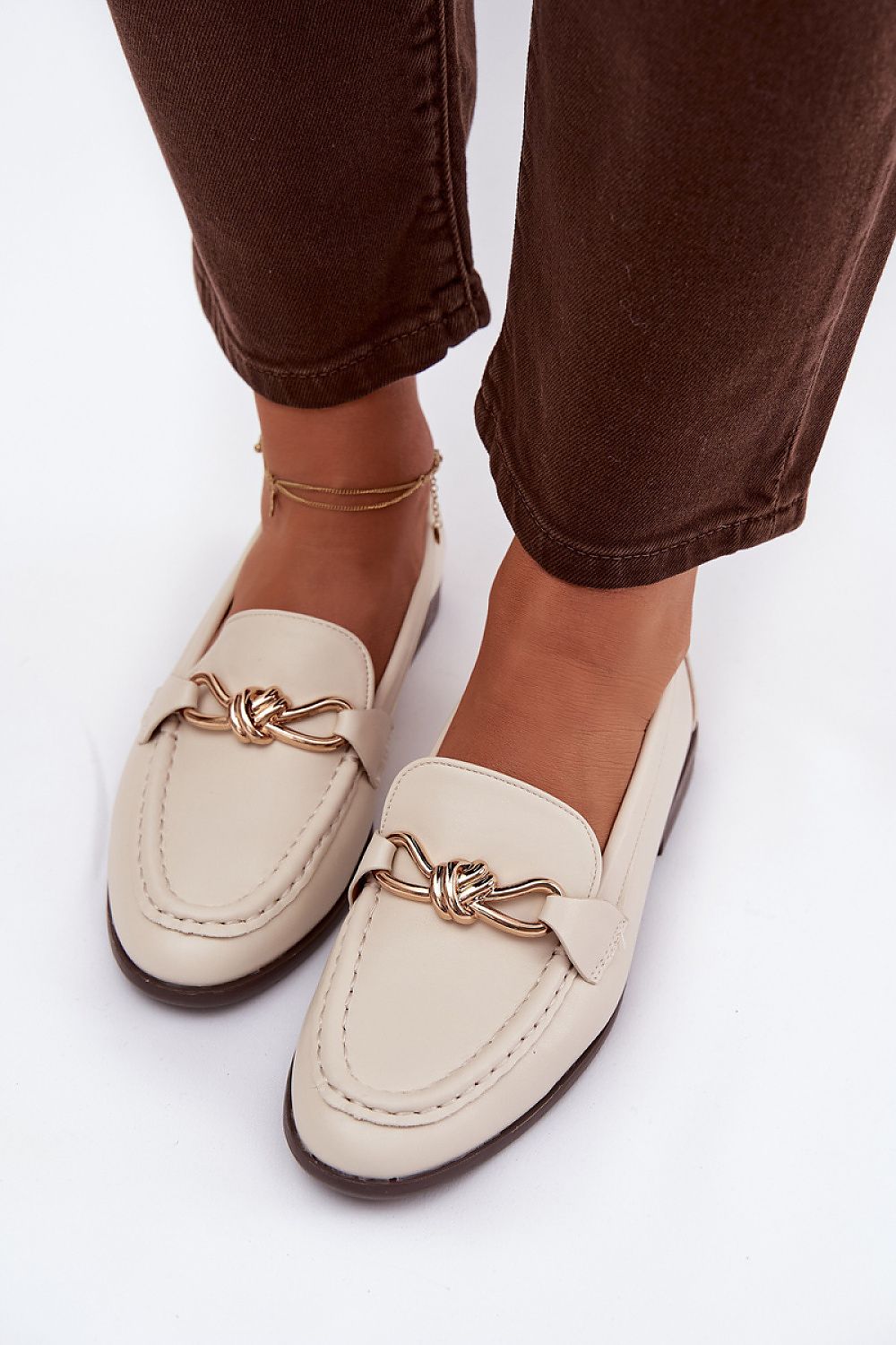 Beige loafers with gold accents worn with brown pants on a white background