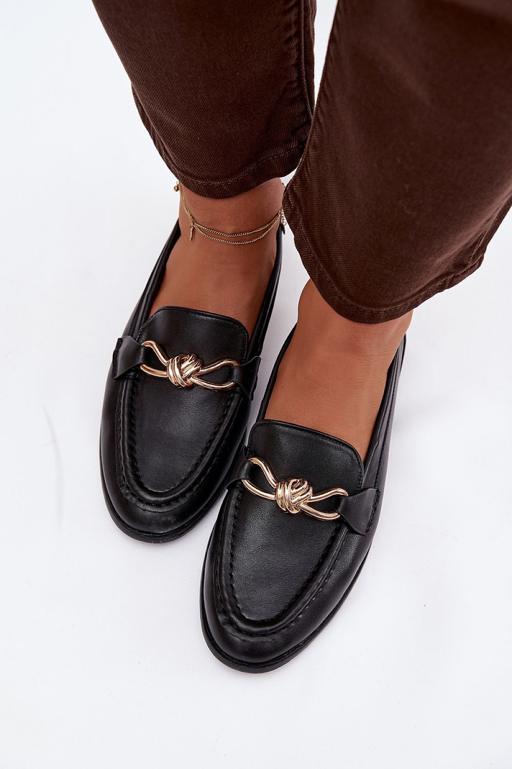 Black loafers with gold accents worn with brown pants on a white background